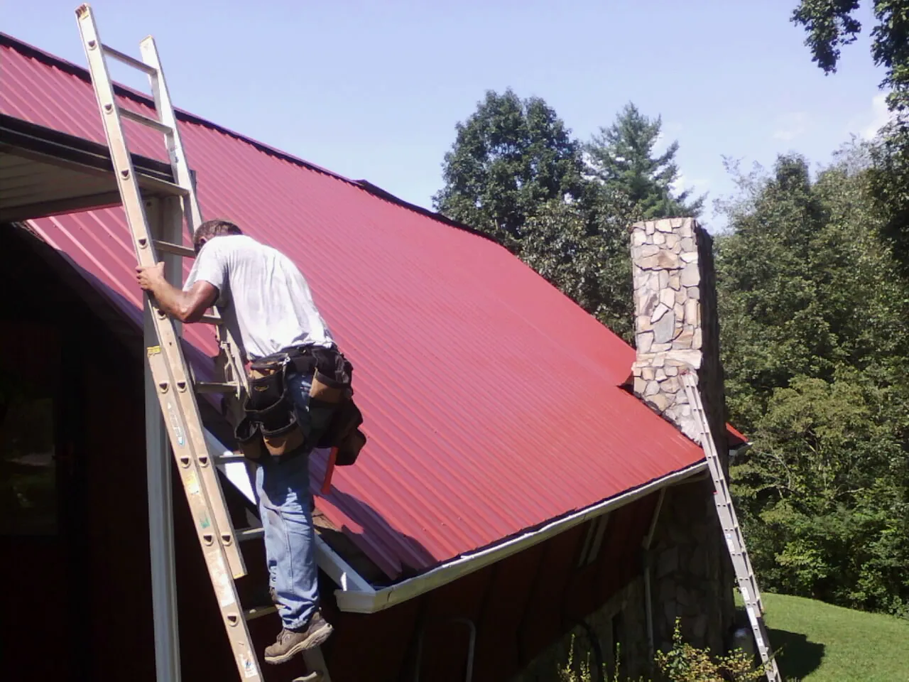 Roofer on a ladder beside a red metal roof and stone chimney — Soffit & Fascia Repair by Roofing Brothers in Altamont